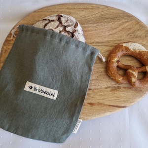 Könnte beinhalten: Ein grauer Brotbeutel aus Leinen mit dem Text "Brotbeutel" auf einem weißen Etikett, neben einem Brotlaib und einer Brezel auf einem Holzschneidebrett. Der Brotbeutel ist teilweise geöffnet und zeigt die Oberseite eines Brotlaibs.