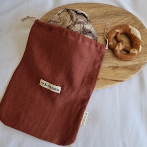 Könnte beinhalten: Ein rostfarbener Brotbeutel aus Leinen mit dem Text "Brotbeutel" auf einem kleinen Etikett. Ein Brotlaib und eine Brezel liegen auf einem Holzschneidebrett. Der Beutel ist offen und zeigt einen Teil des Brotes.