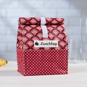 Op de afbeelding: Rode lunchtas met een wit stippenpatroon aan de onderkant en een zwart-wit geometrisch patroon aan de bovenkant. De tas heeft een witte band en een label met de tekst "Lunchbag" met een rood appelpictogram.
