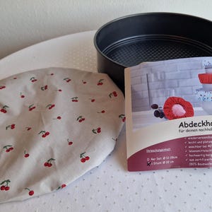 Könnte beinhalten: Eine schwarze runde Backform, eine beige Stoff-Schüsselhaube mit rotem Kirschmuster und eine Produktverpackung mit dem Text "Abdeckhauben". Die Verpackung zeigt Bilder von Schüsselhauben und hat den Text "für deinen nachhaltigen Alltag".