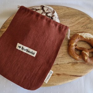 Könnte beinhalten: Ein rostfarbener Brotbeutel mit dem Text "Brotbeutel" auf einem weißen Etikett, neben einem Brotlaib und einer Brezel auf einem Holzschneidebrett. Der Beutel besteht aus einem weichen, strukturierten Stoff und hat einen Kordelzug.