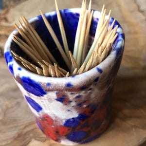 May include: A ceramic toothpick holder with a blue, red, and white splatter paint design. The holder is filled with wooden toothpicks.