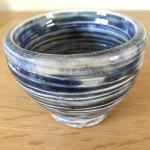 Puede incluir: Un tazón de cerámica blanca con rayas azules y doradas. El tazón tiene una forma redondeada y un borde ancho.