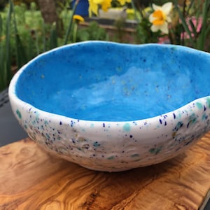 May include: A white ceramic bowl with a blue glaze and speckled white, blue, and green accents. The bowl is sitting on a wooden surface.