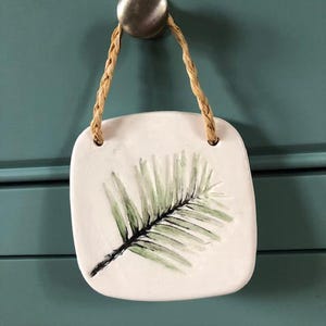 Puede incluir: Un colgante de pared de cerámica blanca con un diseño de hoja de palma verde. El colgante está unido a una cuerda de yute natural.