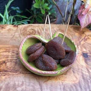 Puede incluir: Un cuenco de cerámica verde con borde rosa lleno de albaricoques secos y palillos. El cuenco está sobre una tabla de cortar de madera con un patrón de vetas naturales. Los albaricoques son de color marrón oscuro.