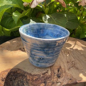 May include: A ceramic bowl featuring a glossy finish with swirling patterns of deep blue, light blue, and white. The bowl has a tapered shape with visible horizontal ridges along the exterior surface and a smooth, rounded rim at the top.