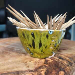 Puede incluir: Un pequeño cuenco de cerámica amarillo y verde con un esmalte moteado, lleno de palillos de dientes de madera.