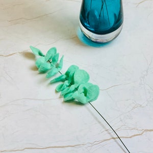 May include: A teal blue glass vase with a single sprig of light green felt eucalyptus leaves. The vase is sitting on a white marble surface.