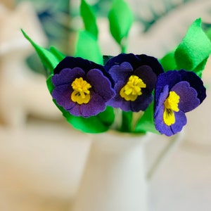 May include: Three purple felt flowers with yellow centres in a white ceramic jug. The flowers are arranged in a bouquet.