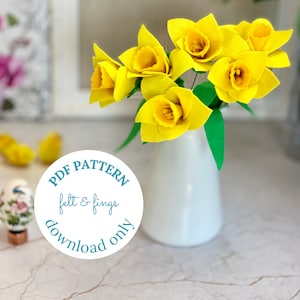 Op de afbeelding: Een witte keramische vaas met een boeket gele vilt narcissen. Een ronde sticker met de tekst "PDF PATTERN felt & fings download only" is op de voorgrond.