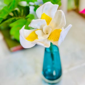 May include: A single white felt flower with yellow accents in a blue glass vase. The flower is a felt craft.