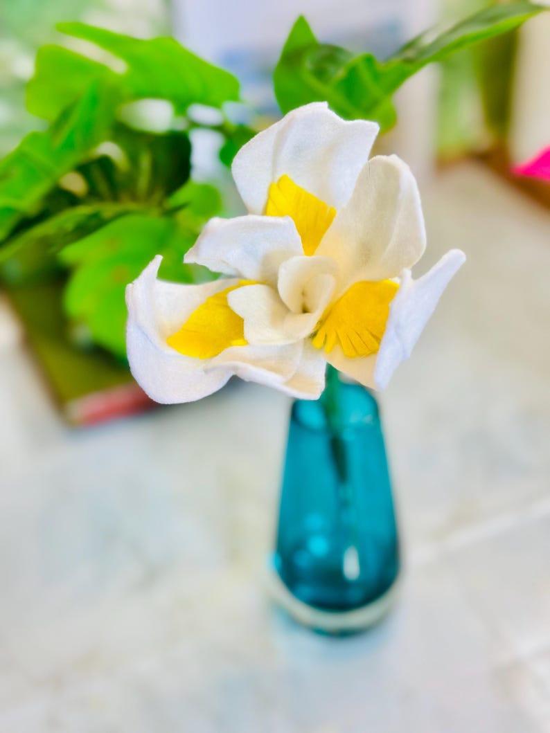 May include: A single white felt flower with yellow accents in a blue glass vase. The flower is a felt craft.