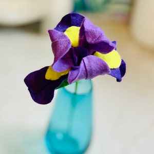 May include: A purple and yellow felt flower in a blue glass vase. The flower is a single bloom with a yellow center and purple petals.
