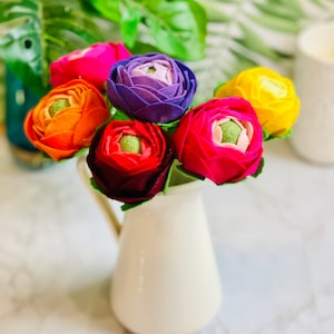 May include: A bouquet of six felt flowers in a white jug. The flowers are in shades of pink, red, orange, yellow, and purple.