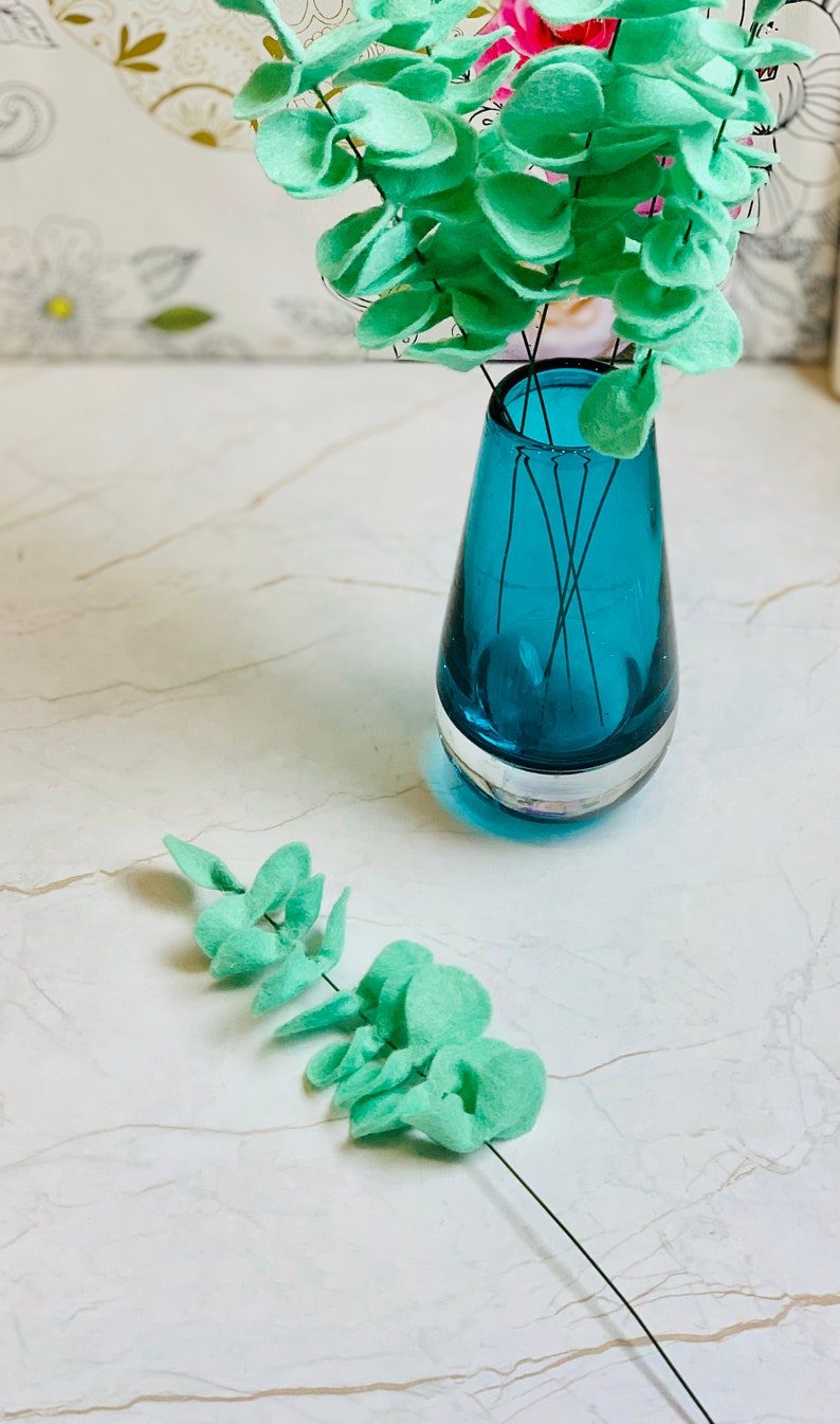 May include: A teal blue glass vase with a single sprig of light green felt eucalyptus leaves. The vase is sitting on a white marble surface.