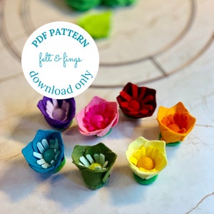 May include: Seven felt flower embellishments in various colors, including purple, pink, red, orange, yellow, green, and blue. The flowers are arranged on a white surface. A white circle with blue text reads "PDF PATTERN felt & fings download only".