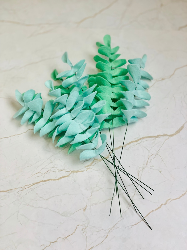 May include: A bunch of faux eucalyptus stems with green and light blue leaves. The stems are made of wire and are ready to be used in a craft project.