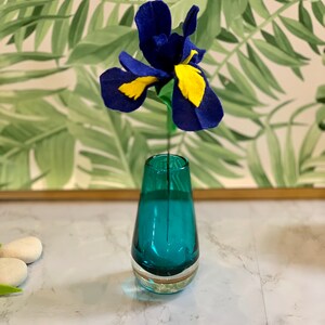 May include: A blue and yellow felt flower in a teal glass vase. The vase is on a white marble surface.