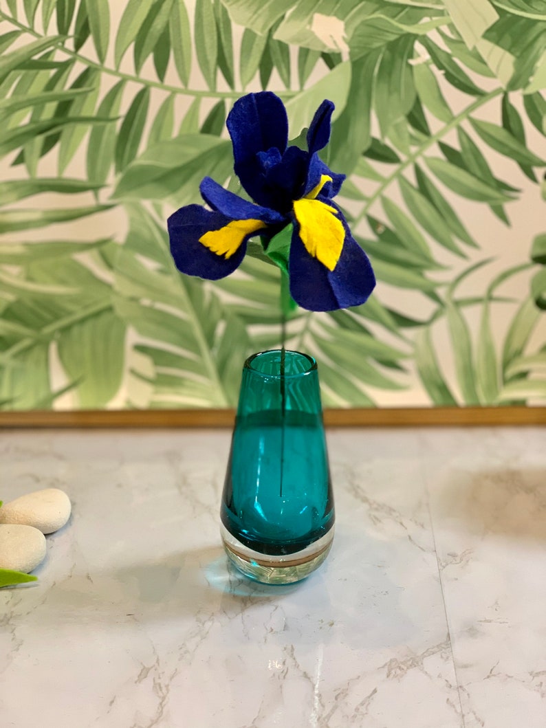 May include: A blue and yellow felt flower in a teal glass vase. The vase is on a white marble surface.