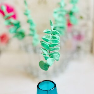 May include: A single sprig of light green faux eucalyptus leaves is suspended above a blue glass vase.