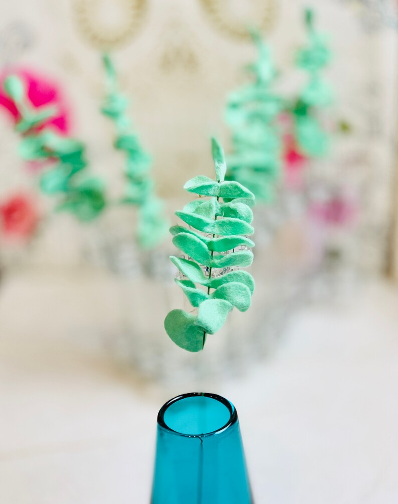 May include: A single sprig of light green faux eucalyptus leaves is suspended above a blue glass vase.