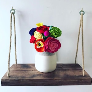 Könnte beinhalten: Ein Holzregal mit Seilhängern, auf dem eine weiße Vase mit einem bunten Filzblumenarrangement steht. Die Blumen sind rot, pink, gelb, grün und blau.