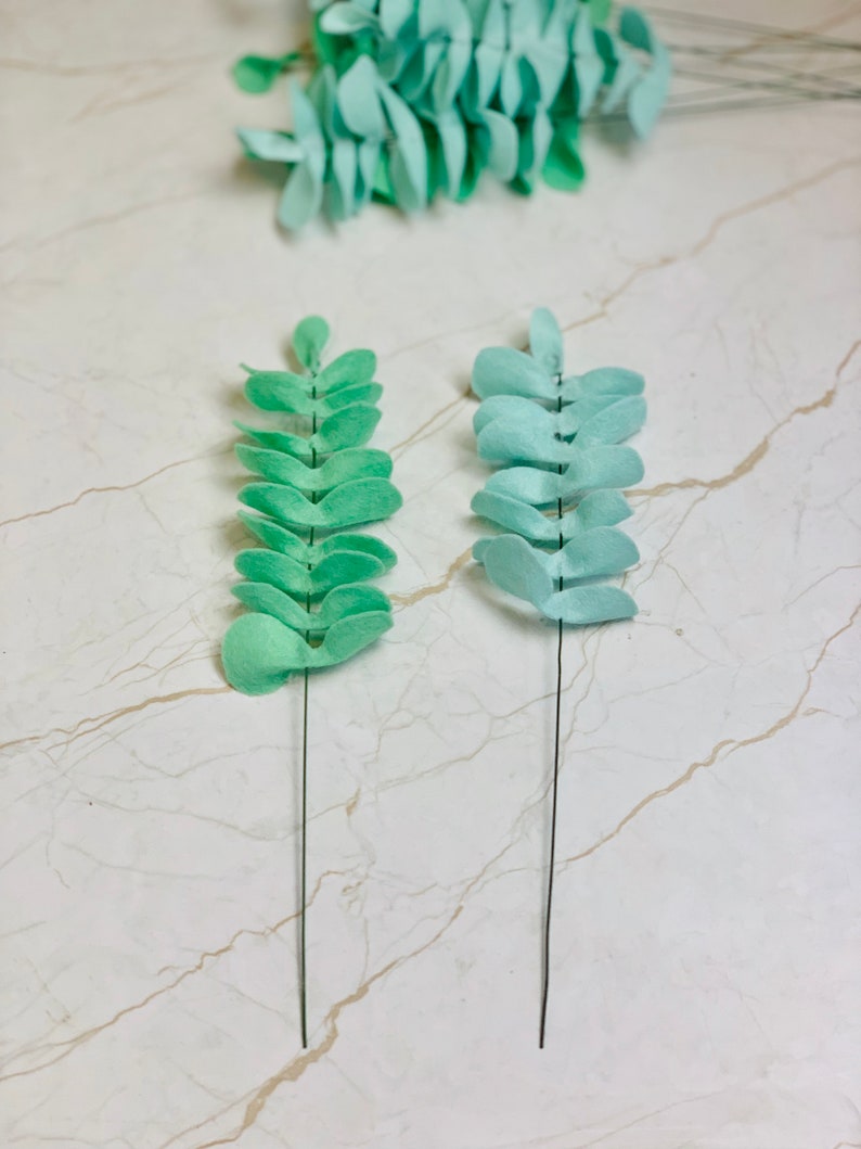 May include: Two sprigs of faux greenery with light green felt leaves attached to thin wire stems. The leaves are arranged in a staggered pattern.