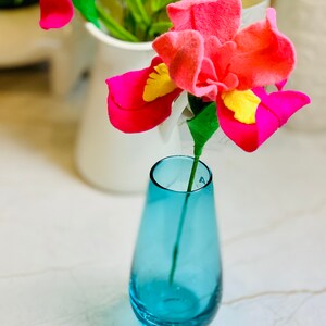 May include: A single pink felt flower with yellow accents in a blue glass vase on a white marble surface.