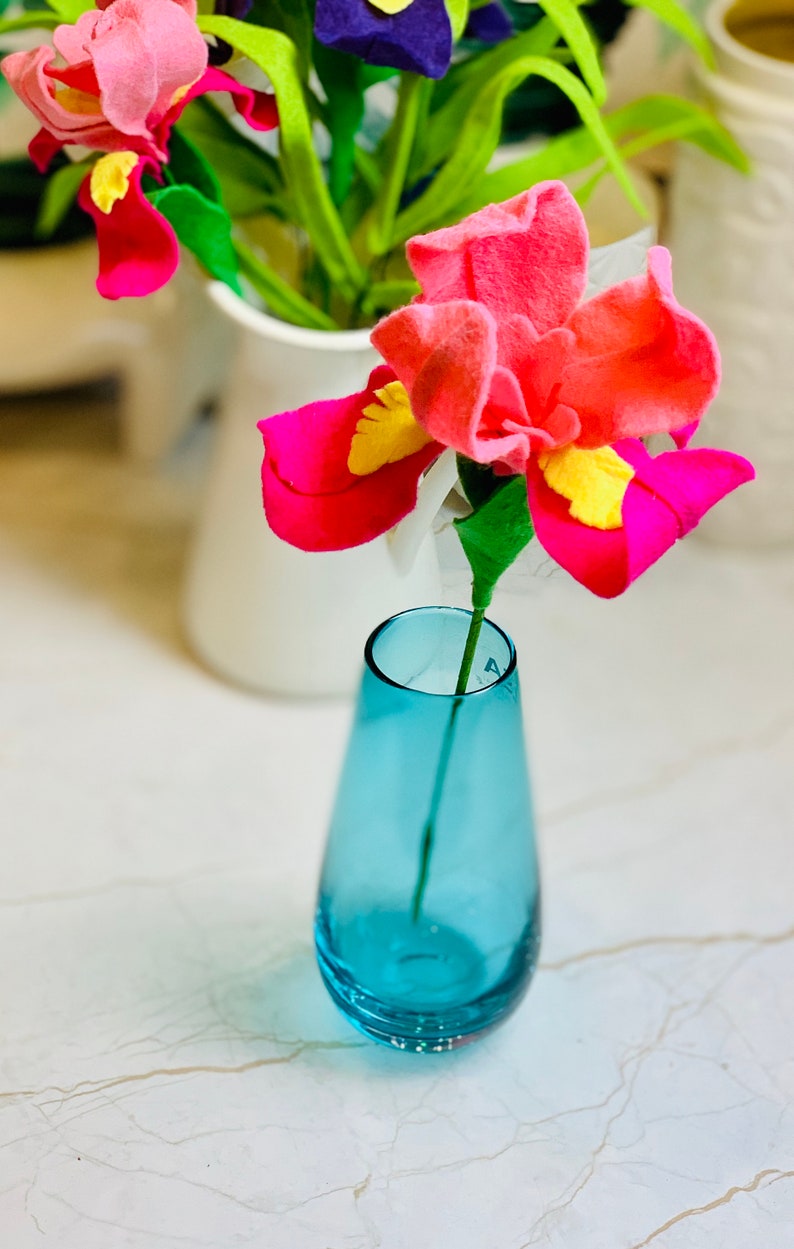 May include: A single pink felt flower with yellow accents in a blue glass vase on a white marble surface.