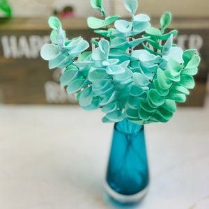 May include: A bouquet of light blue and green felt eucalyptus leaves in a teal blue vase.