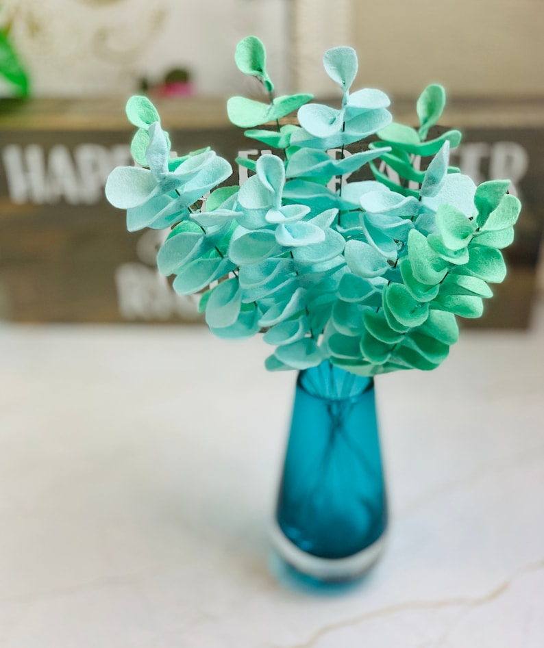 May include: A bouquet of light blue and green felt eucalyptus leaves in a teal blue vase.