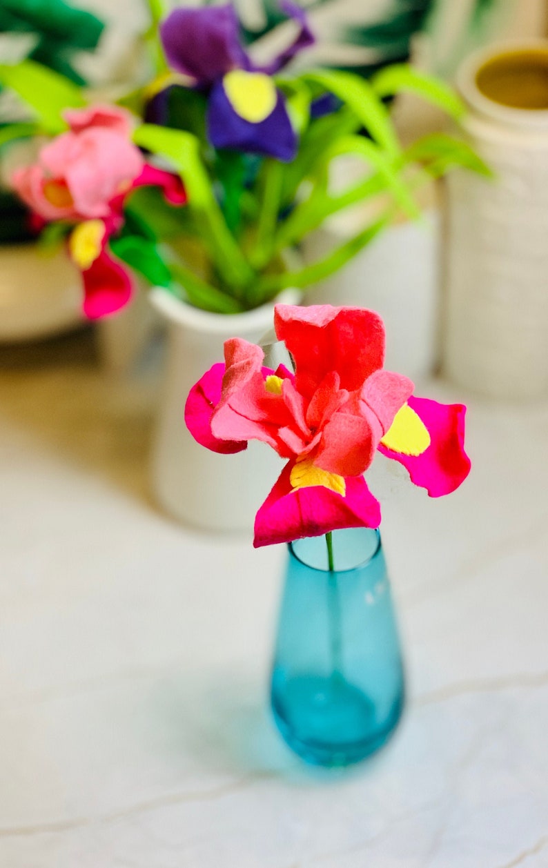 May include: A single pink and yellow felt flower in a blue glass vase. The flower is a felt craft and is sitting on a white surface.