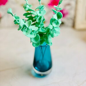 May include: A bouquet of light green felt eucalyptus leaves in a teal blue glass vase.