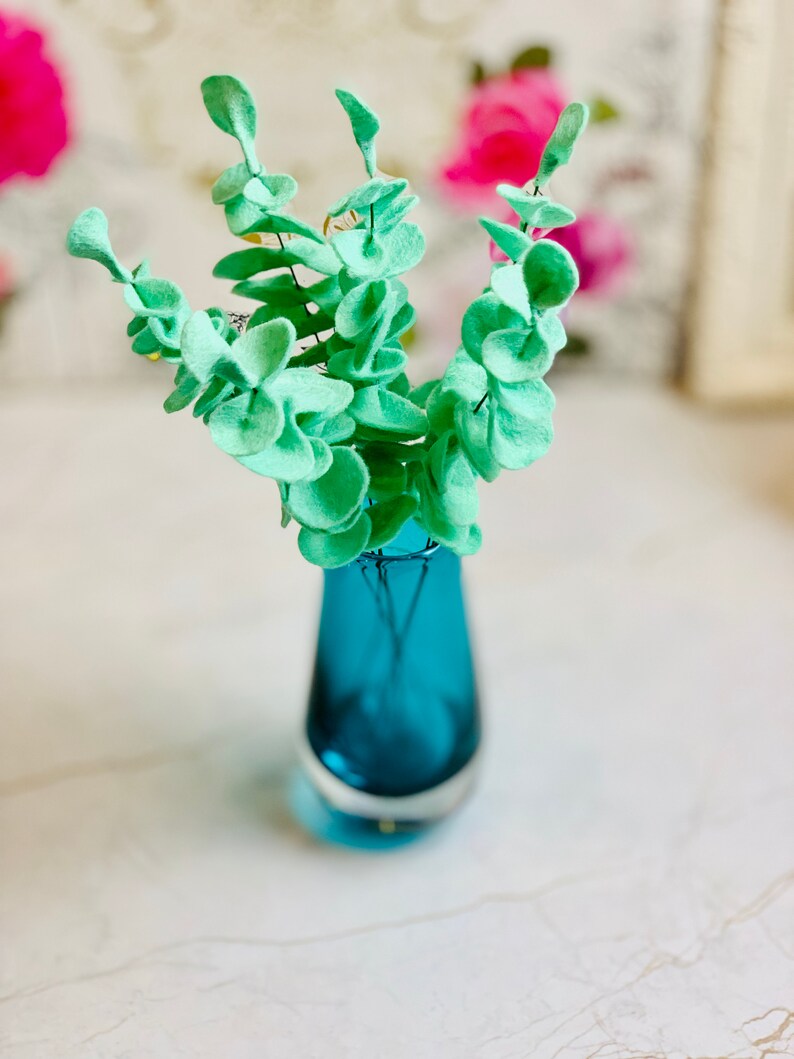 May include: A bouquet of light green felt eucalyptus leaves in a teal blue glass vase.