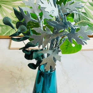 May include: A teal glass vase with a light blue felt flower arrangement. The flowers are made of felt and have a delicate, leafy design.
