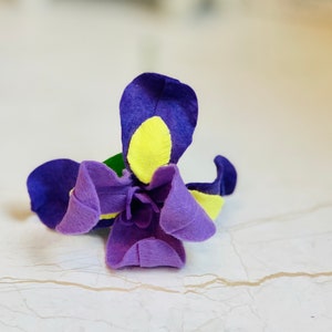 May include: A purple felt flower with yellow accents. The flower is made of felt and has a three-dimensional shape.