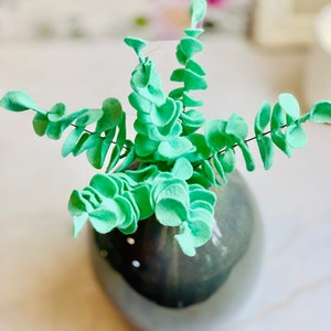May include: A vase filled with a faux eucalyptus plant made of felt. The leaves are a light green color and are arranged in a spiral pattern. The vase is a dark gray color and has a smooth, glossy finish.