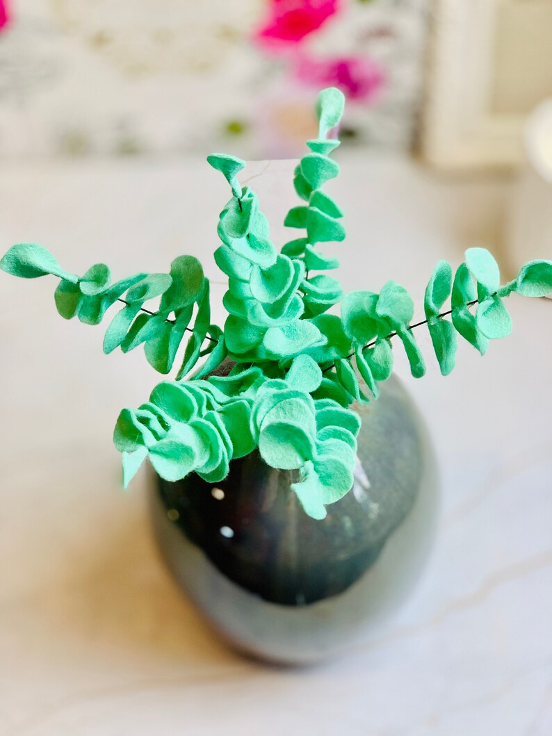 May include: A vase filled with a faux eucalyptus plant made of felt. The leaves are a light green color and are arranged in a spiral pattern. The vase is a dark gray color and has a smooth, glossy finish.