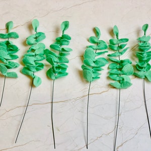 May include: Seven sprigs of faux eucalyptus leaves, each with a wire stem, are arranged on a white surface. The leaves are a light green color and are made of felt.
