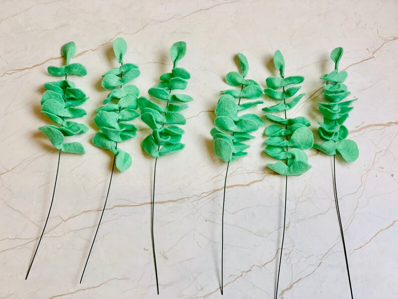 May include: Seven sprigs of faux eucalyptus leaves, each with a wire stem, are arranged on a white surface. The leaves are a light green color and are made of felt.