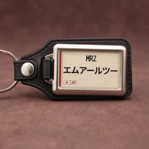 Op de afbeelding: Een zwarte leren sleutelhanger met een rechthoekig metalen frame en een witte inzet. De inzet toont de tekst MR2, de Japanse tekst エムアールツー en een klein rood vierkant met de letters JP. Een zilveren metalen ring is aan het leer bevestigd.