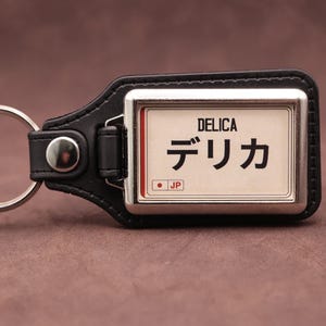 Könnte beinhalten: Ein schwarzer Lederschlüsselanhänger mit einem rechteckigen Metallrahmen und einem weißen Einsatz. Der Einsatz zeigt den Text DELICA in Schwarz, die japanischen Schriftzeichen デリカ und ein kleines rotes Quadrat mit den Buchstaben JP neben einer japanischen Flagge.