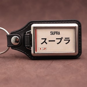 Puede incluir: Un llavero de cuero negro con un marco metálico rectangular y una inserción blanca. La inserción muestra el texto SUPRA en negro, los caracteres japoneses スープラ y un pequeño cuadrado rojo con las letras JP y un punto.