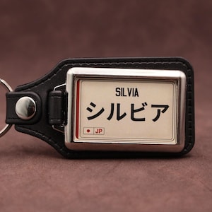 Pode incluir: Um porta-chaves de couro preto com uma moldura metálica retangular e uma inserção branca. A inserção apresenta o texto SILVIA em preto, o texto japonês シルビア e um pequeno círculo vermelho com as letras JP numa caixa branca.