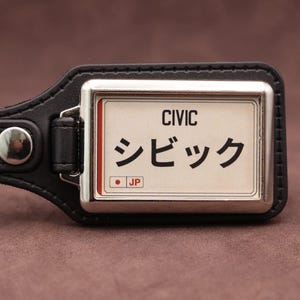 Honda Civic Japanese plate medallion keyring with black leatherette fob, chrome frame and cream insert on a brown suede background.