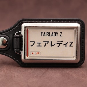 May include: A black leather keyring featuring a rectangular metal plate with a white background. The plate displays the text FAIRLADY Z, the Japanese characters フェアレディZ, and a small red circle with the letters JP in a white box.