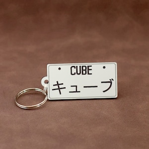 May include: White keychain with a black license plate design. The plate says "CUBE" and "キューブ" in Japanese characters.