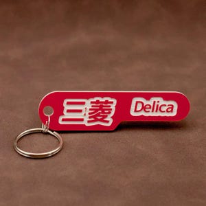 Op de afbeelding: Rode metalen sleutelhanger met het woord "Delica" en Japanse karakters in wit. De sleutelhanger heeft een zilveren ring.