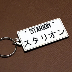 Pode incluir: Chaveiro branco com uma etiqueta retangular preta que diz "STARION" e "スタリオン" em japonês. O chaveiro tem um anel de metal prateado.